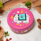 A round, pink spice box with a hand-painted white elephant design and colorful floral motifs. The box is placed on a white surface with a plant and a teddy bear in the background.